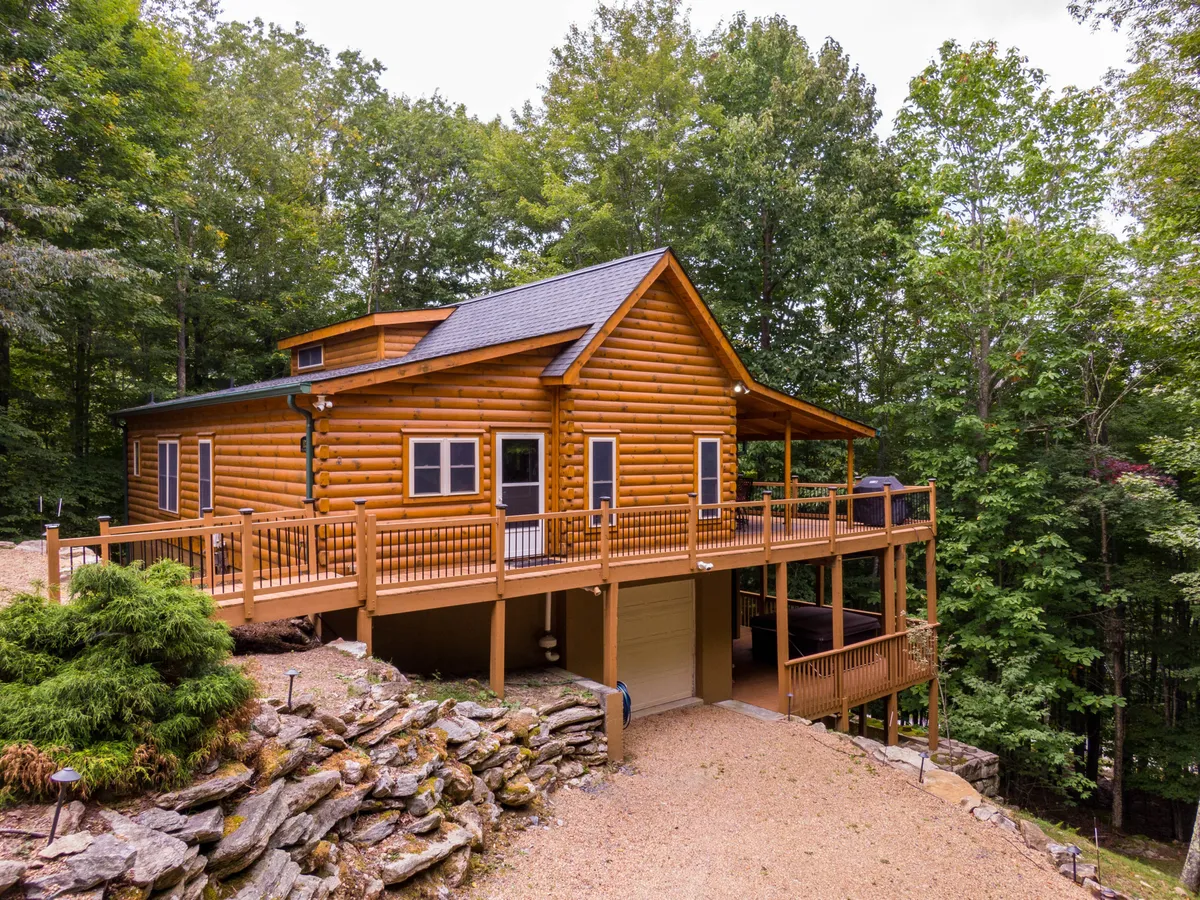 A welcoming log cabin with classic mountain architecture is nestled among lush trees, featuring a spacious deck that invites guests to gather, enjoy barbecues, or set out on nature walks. The surrounding forest hints at the endless outdoor adventures available nearby.