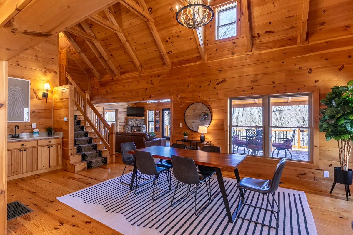 A cozy dining area with a large wooden table under a statement chandelier, adjacent to a staircase and surrounded by wood-paneled walls and generous windows. The abundance of natural light and comfortable seating makes it ideal for shared meals and festive gatherings, while the setting hints at both intimacy and celebration.