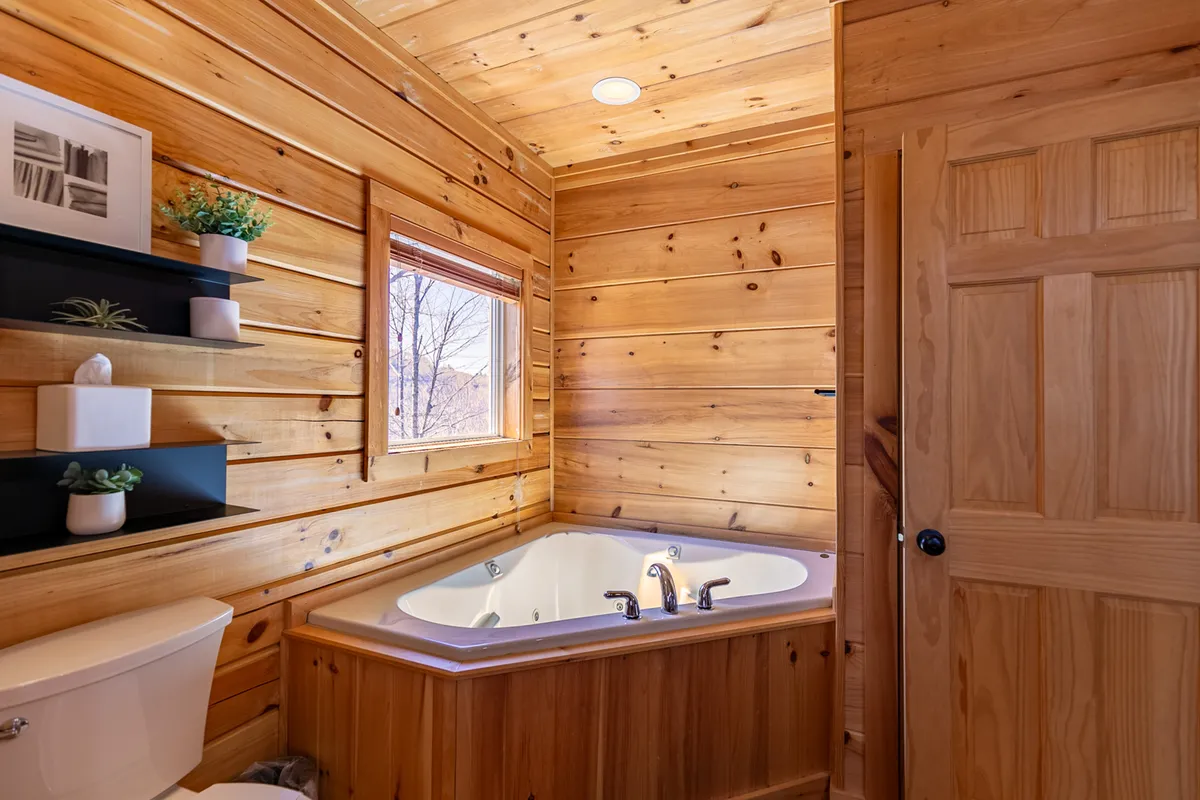 A serene bathroom featuring a jacuzzi bathtub set against warm wooden paneling, with a window offering natural light and a scenic view. A shelf with decor and soothing design elements evoke a spa-like ambiance.