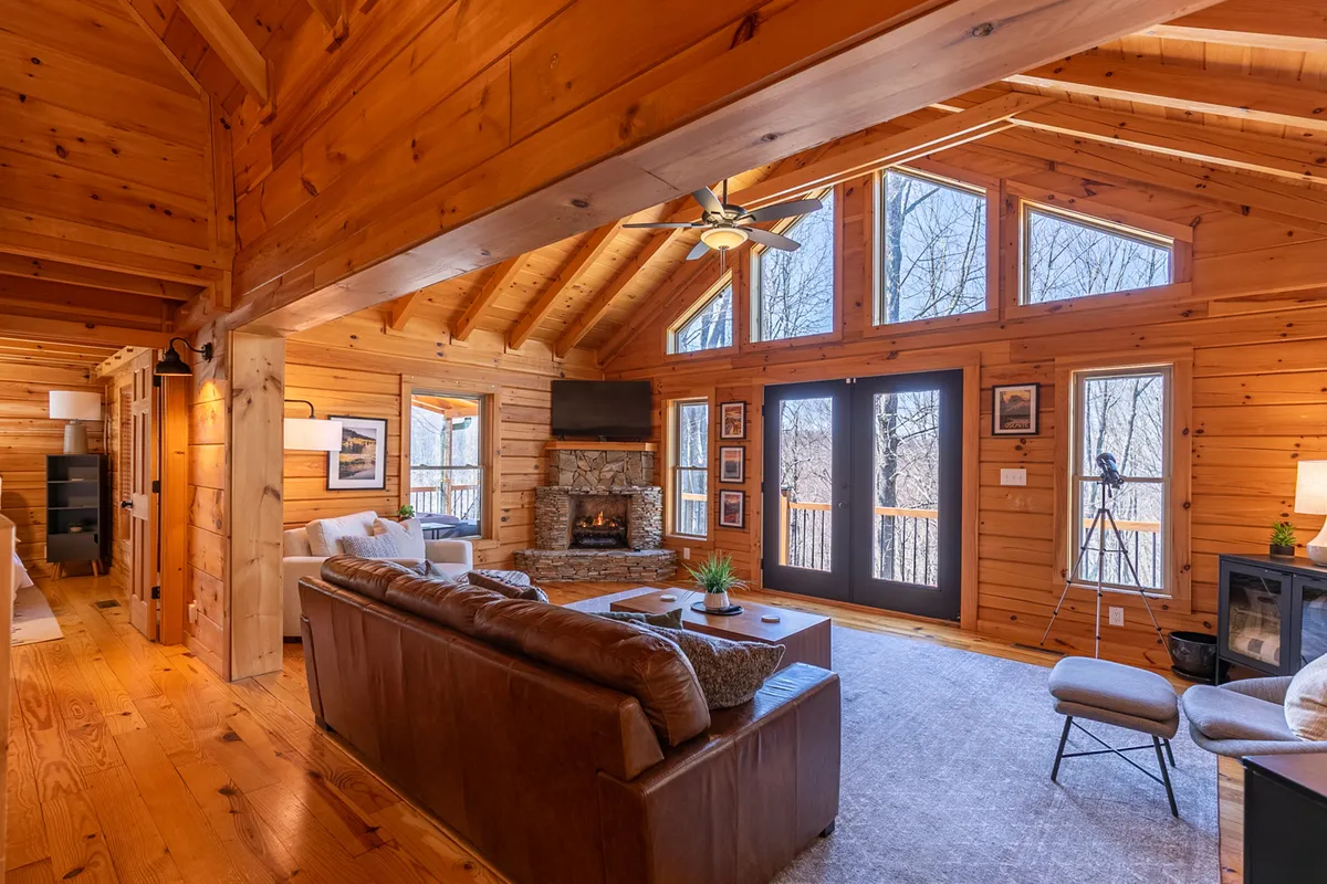 A warm, inviting great room featuring a stone gas fireplace, comfortable leather sofa, and expansive wooden beams and paneling. Large windows offer scenic views of the surrounding forest, filling the space with natural light and creating a connection to nature. The room is designed for relaxation and gathering, with plush seating ideal for unwinding after a day of mountain adventure.