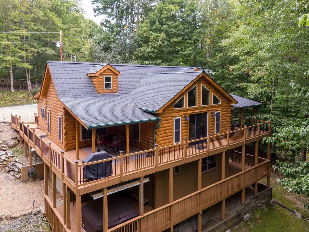 This image showcases the exterior of The Mountainbriar Lodge—a charming log cabin-style home nestled among lush trees. The spacious deck wraps around the house, adorned with comfortable seating for outdoor dining or relaxing while taking in the surrounding natural beauty. The scene is bathed in the warm glow of sunset, suggesting a tranquil and cozy mountain retreat.