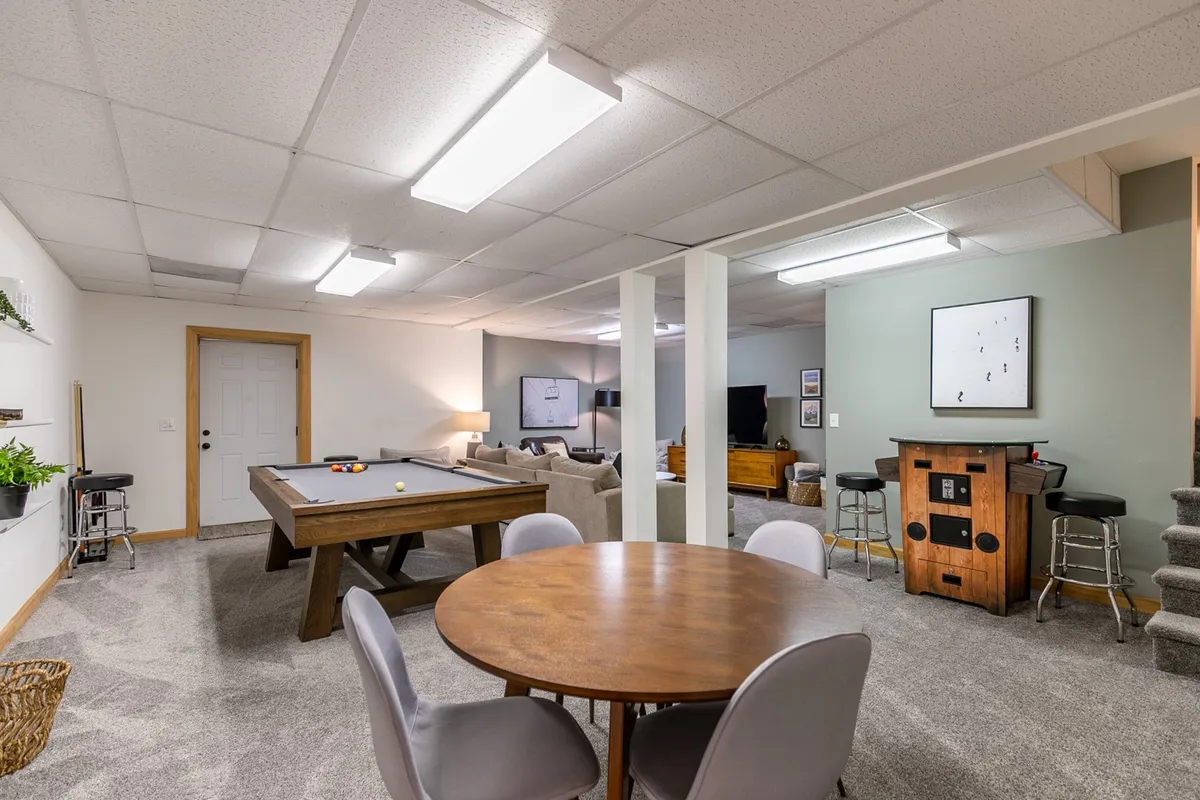 A wide view of the entertainment area showcasing a pool table, a round dining/game table, a dedicated media area with a TV, and a stylish bar station. The space is arranged to support a variety of activities from games and socializing to casual dining and movie marathons.
