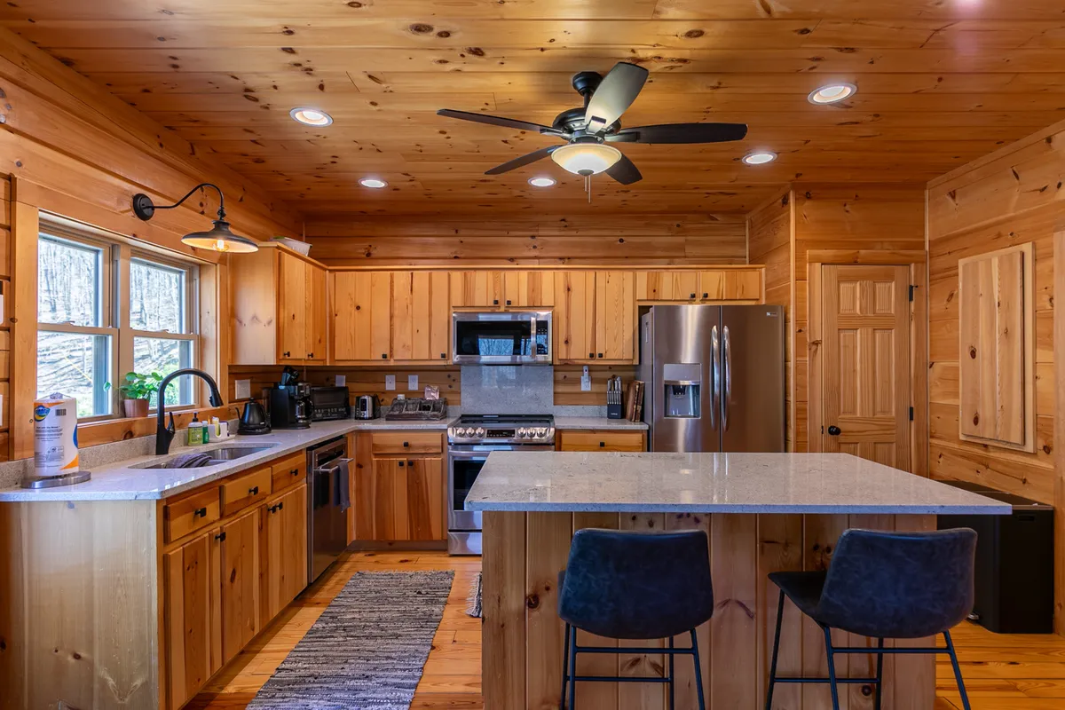 A chef-ready kitchen with a central island, stainless steel appliances, and wooden cabinetry. Large windows provide ample daylight and forest views, making the kitchen both functional and scenic. The kitchen island invites guests to gather, cook, and share stories while preparing meals together.