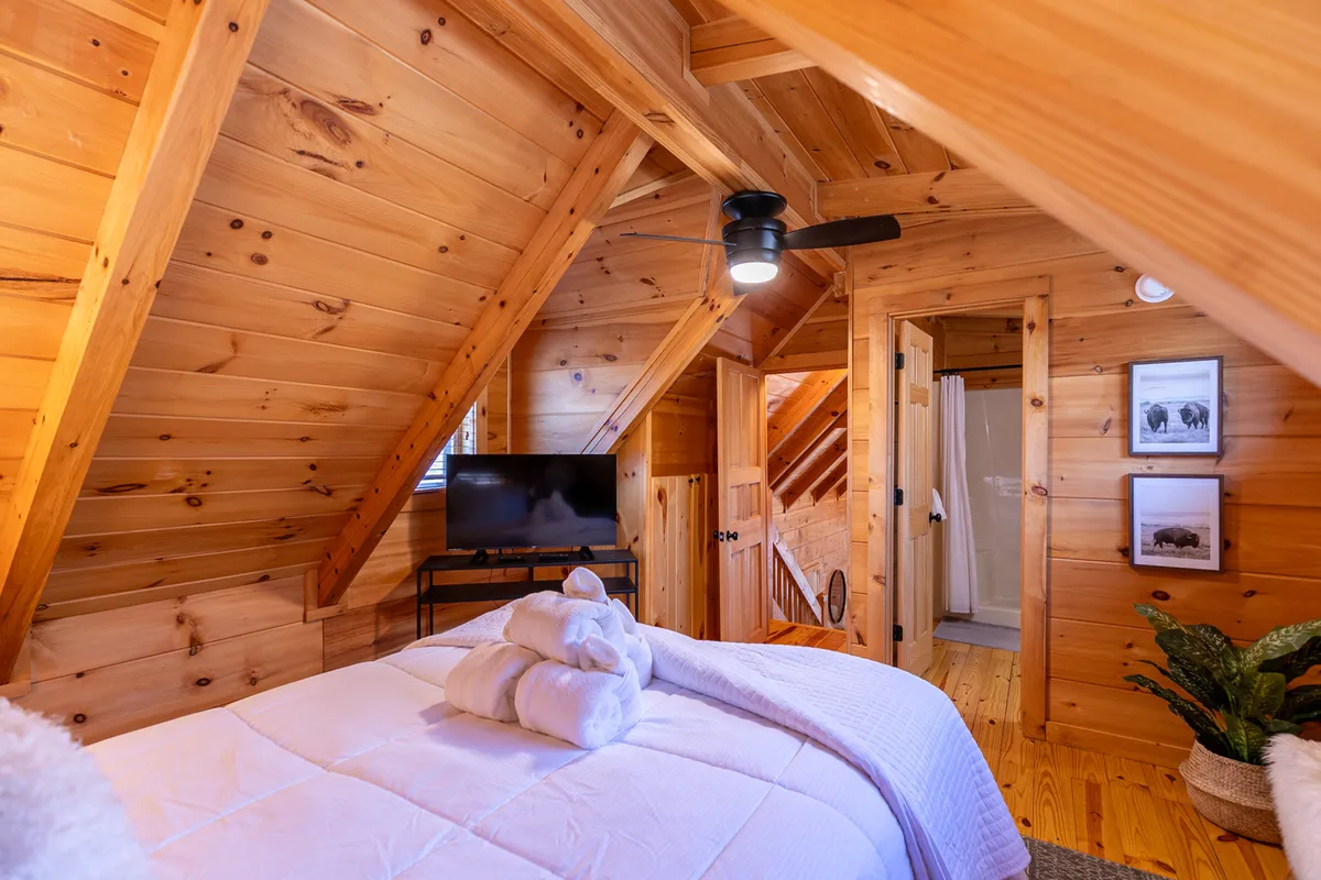 A spacious bedroom with a plush king bed draped in soft linens, tall wooden ceilings with exposed beams, a wall-mounted television, and tasteful artwork. A potted plant adds a touch of natural calm. The ambiance is relaxed and inviting, ideal for winding down after a day outdoors.