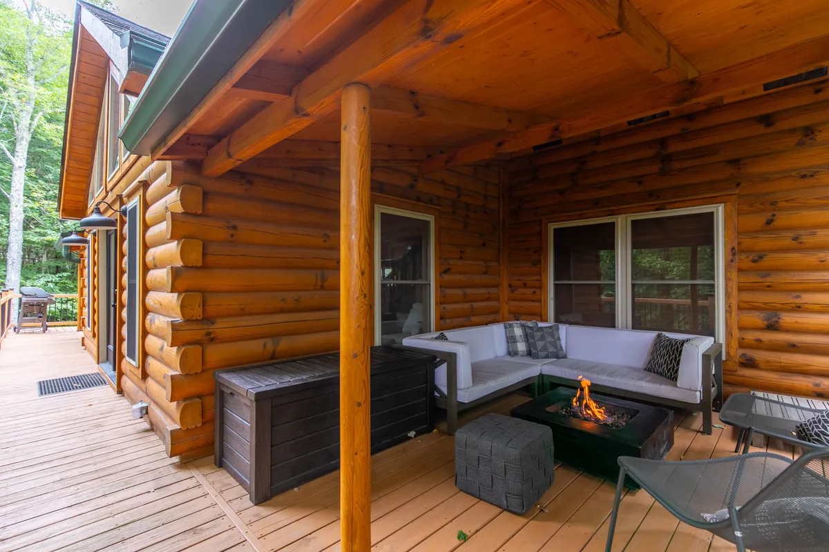 This image captures the inviting outdoor fire pit and seating area on the wooden deck of the log cabin. The scene is set for group gatherings, with comfortable seating for multiple people, and the cabin’s exterior and forest backdrop create a cozy, social atmosphere.