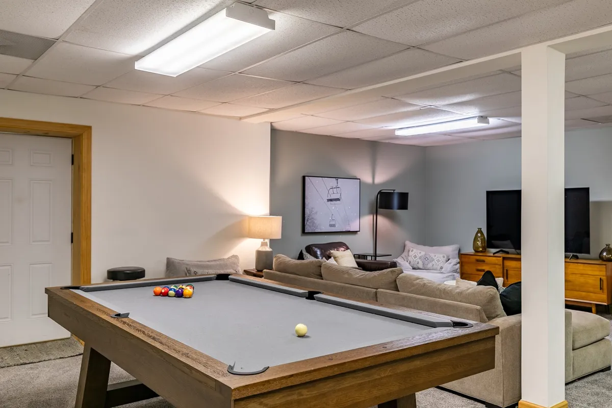 A bright, open game room featuring a stylish pool table at the center, a comfortable sofa nearby, a standing lamp, and a large screen TV mounted on the wall. The layout suggests an area designed for both friendly competitions and relaxed movie nights, with space for gathering and conversation.