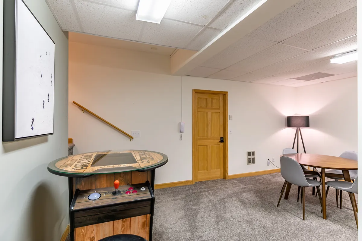 A cozy and engaging area that centers around an arcade game table, with a dining/card table and warm lighting. The scene invites groups to enjoy retro gaming tournaments, board games, and conversations, reinforcing the social and competitive spirit of the space.