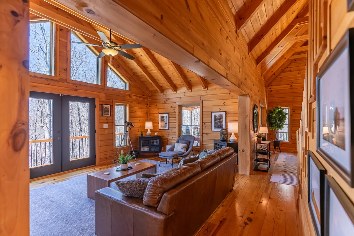 A spacious living area featuring large windows with views of the natural setting, exposed wooden beams, and inviting, comfortable seating arranged around a coffee table. The scene captures both the warmth of the interior and the connection to the outside world.